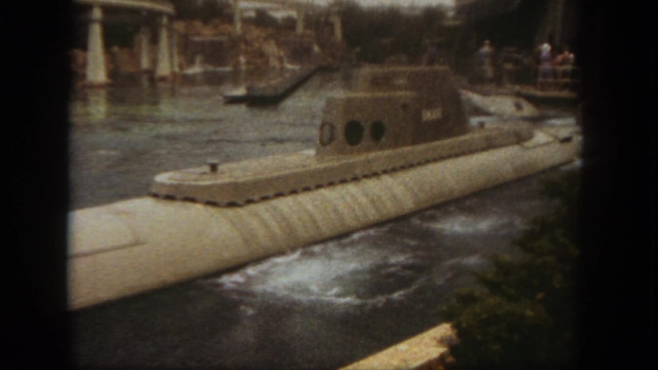 Gray submarine ride vehicle marked SKATE moving through a water channel with guests in the background.