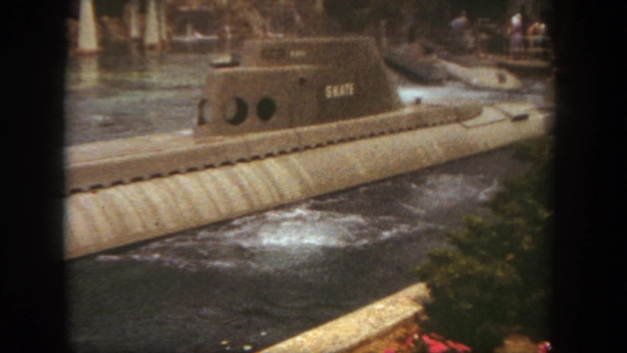 The SKATE submarine continues along the Disneyland waterway, framed by rippling water, foliage, and distant park guests in this 1962 8mm still.