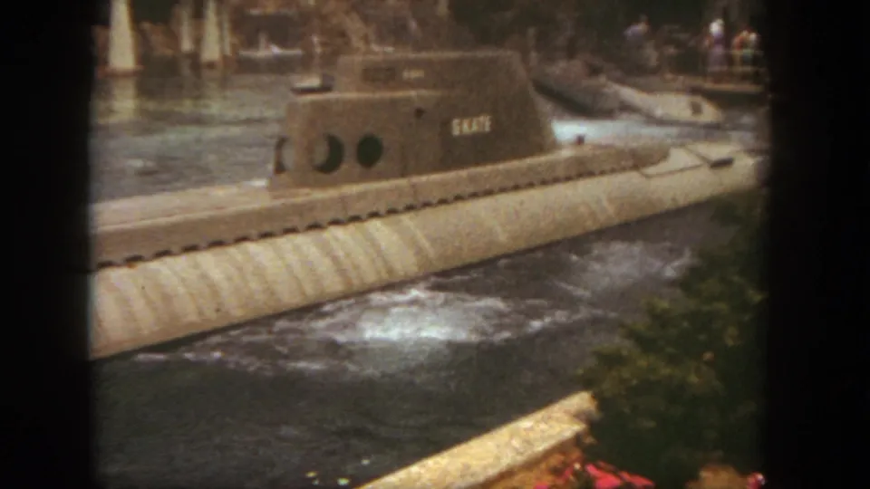The SKATE submarine continues along the Disneyland waterway, framed by rippling water, foliage, and distant park guests in this 1962 8mm still.