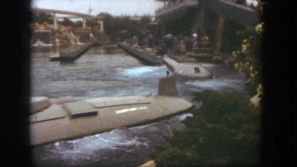 A Disneyland submarine-style ride boat glides past the camera as guests stand near the waterway and bridge structures.