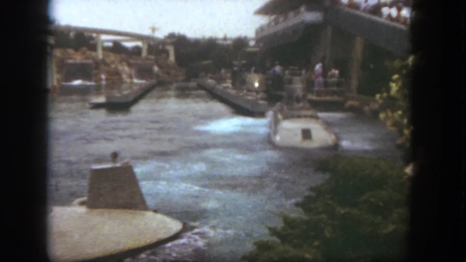 A submarine-style ride boat travels away through the Disneyland waterway, framed by arched bridges, plants, and spectators.