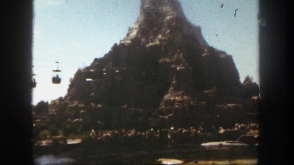 Handheld 8mm Disneyland frame showing the Matterhorn, suspended cable cars, water, boats, and visitors below.