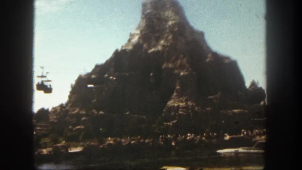 1962 Disneyland Matterhorn view with a cable car entering from the left, visitors below, and boats on the water.