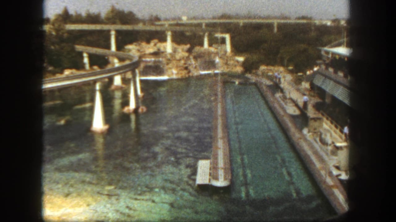 Aerial view over a turquoise water channel, rock landscaping, and elevated tracks.