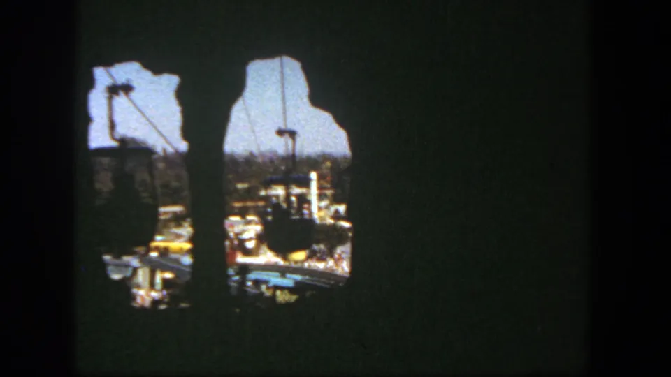 Damaged 8mm frame showing dark gate area with two bright openings revealing cable cars, tracks, and park scenery beyond.