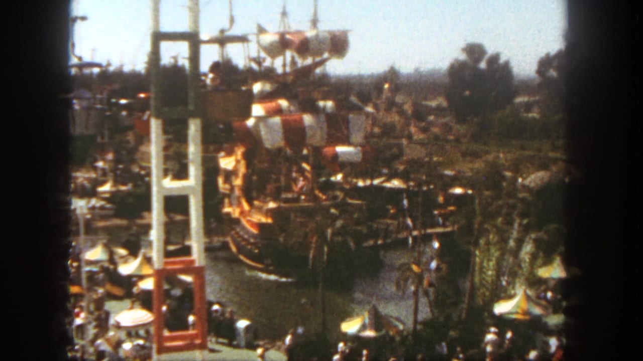 1962 Disneyland home-movie view of a pirate ship ride with red and white sails, tall towers, trees, and visitors below.