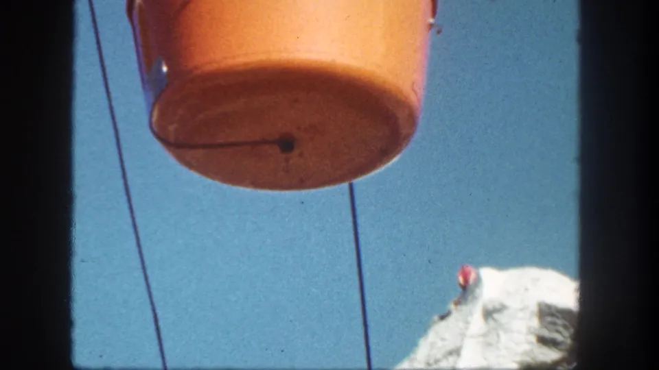 Orange Disneyland Skyride bucket fills the frame as it passes overhead, with cable lines and a pale rock peak lower in view.