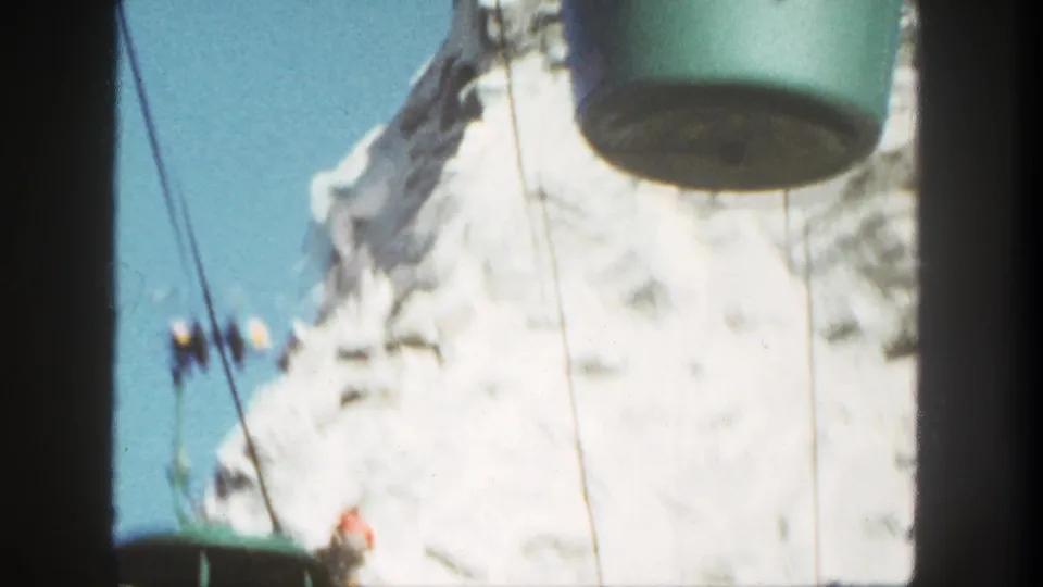 1962 Disneyland frame with a green cableway bucket moving past the snow-covered mountain, cables, and distant climber figures.