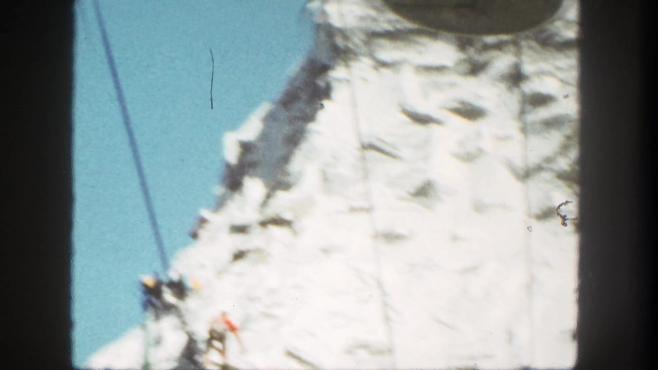 1962 Disneyland home-movie frame showing a bright artificial cliff face, ride cables, blue sky, and small climber figures.