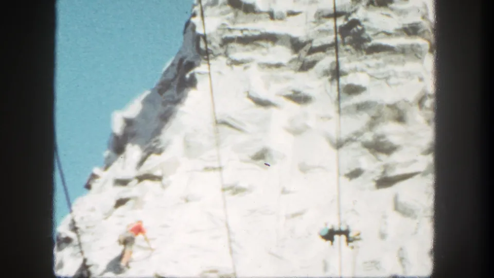 1962 Disneyland home-movie frame showing a red-shirted climber low on the cliff beside visible attraction cables.