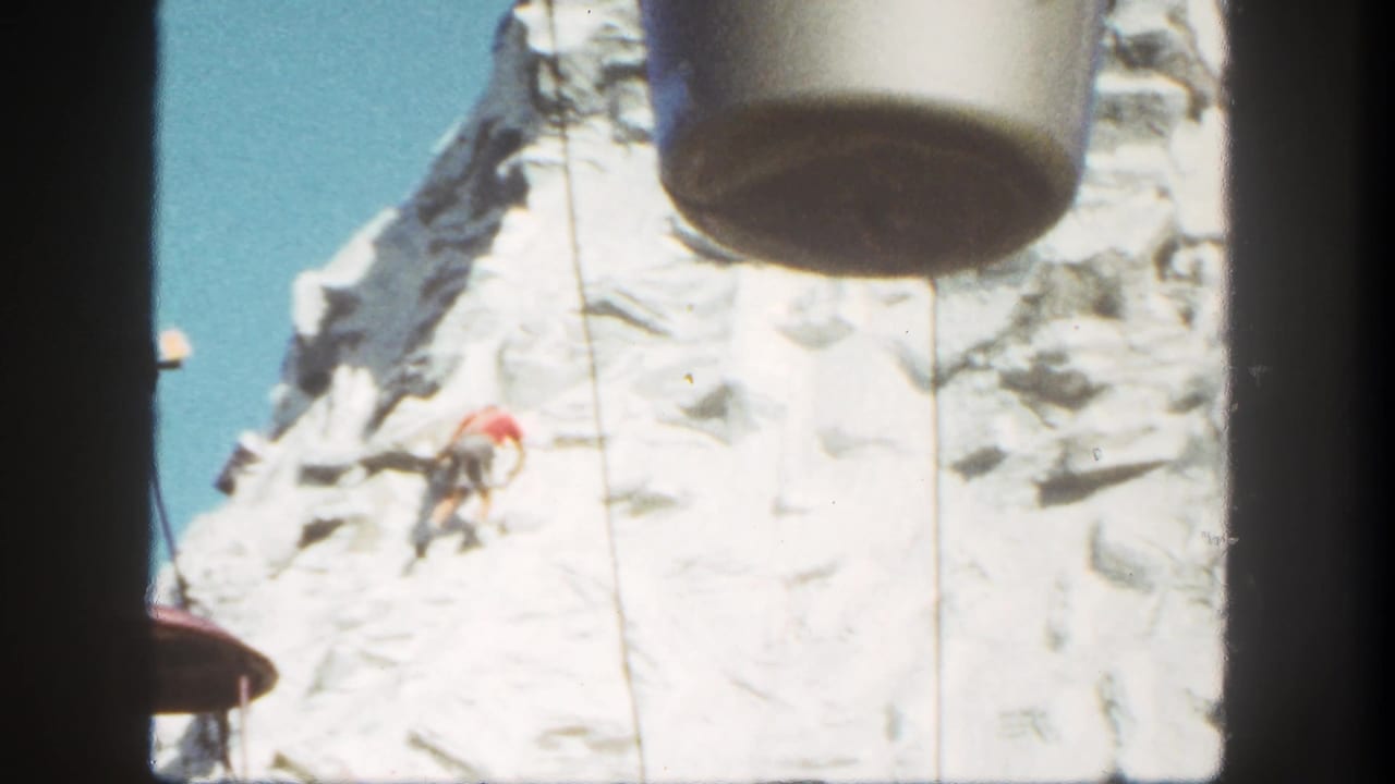 1962 Disneyland view of a red-shirted climber on a snowy artificial mountain beneath a passing gondola bucket.
