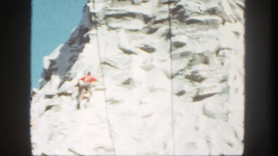 1962 Disneyland footage frames the climber lower on the snowy artificial mountain, with rope lines and film edge visible.