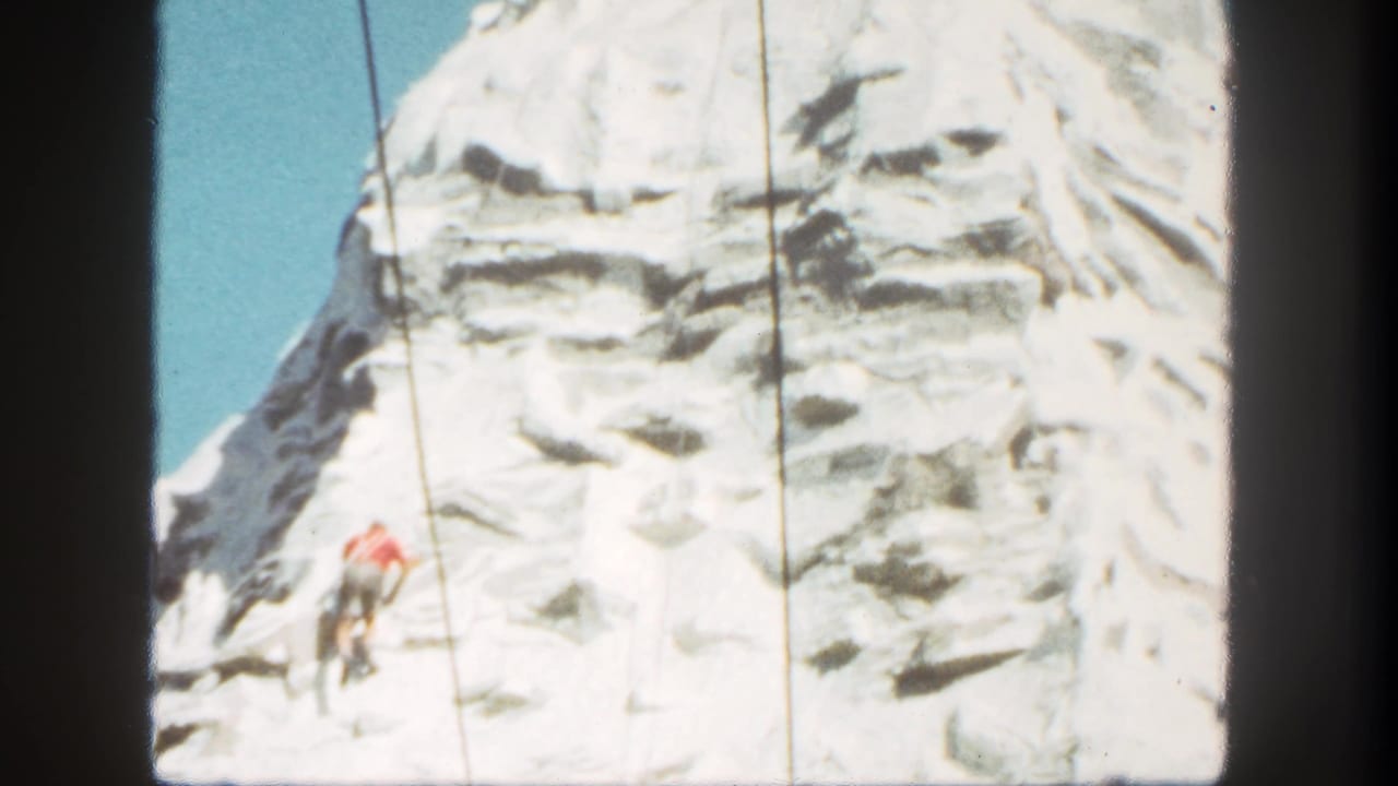 Climber in a red shirt on a white artificial mountain face with ropes against blue sky.