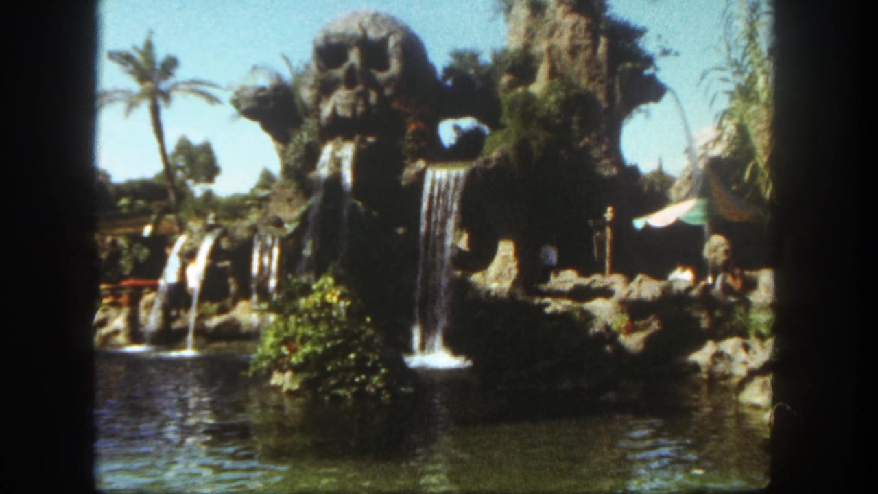 1962 Disneyland home-movie frame showing a tropical attraction with skull-like rockwork, cascading waterfalls, lagoon water, palms, and visitors.