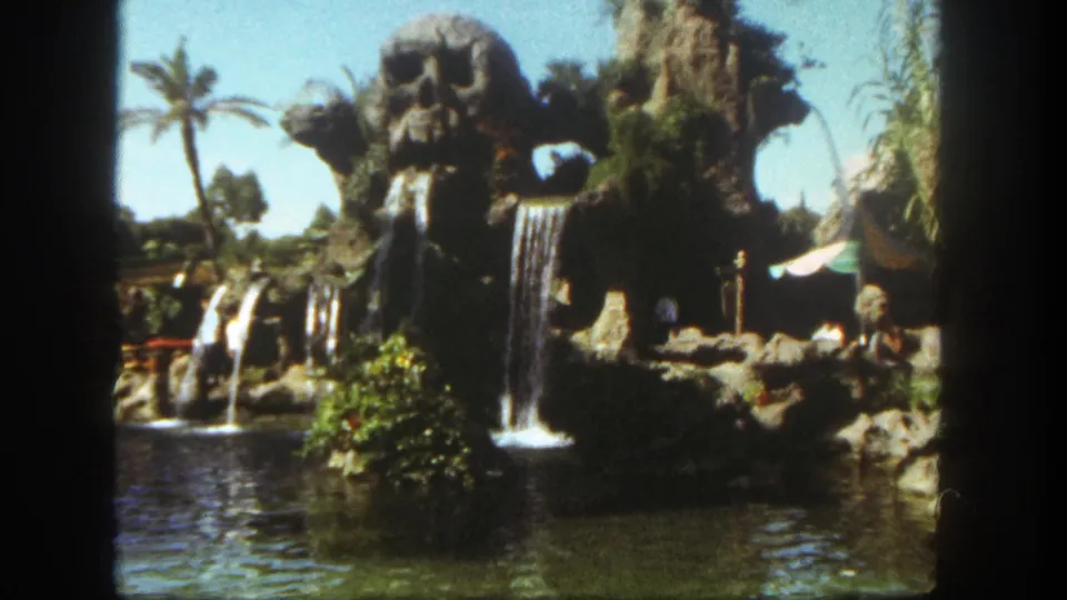 1962 Disneyland home-movie frame showing a tropical attraction with skull-like rockwork, cascading waterfalls, lagoon water, palms, and visitors.