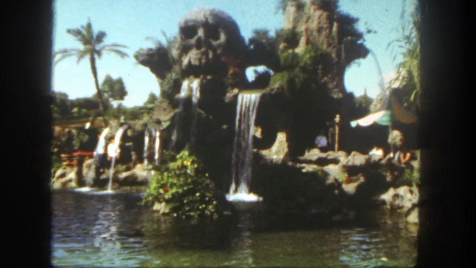 1962 Disneyland home-movie frame of a tropical-themed waterfall attraction with skull-like rockwork, palm tree, lagoon water, and visitors at the edge.