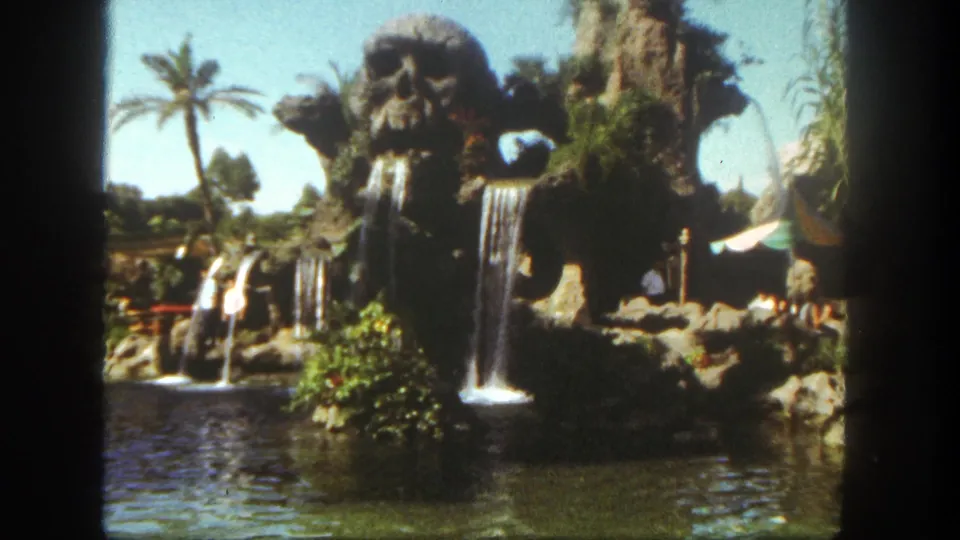 1962 Disneyland home-movie frame showing the skull-like waterfall attraction, lagoon water, palm tree, rockwork, and small visible visitors.
