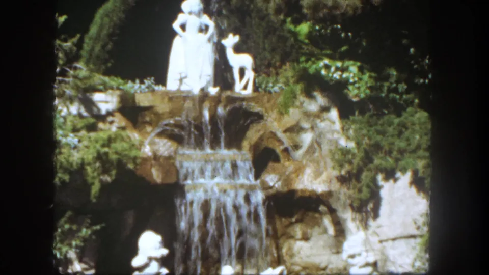 1962 Disneyland home-movie frame showing white Snow White and deer statues above a rock waterfall surrounded by garden planting.