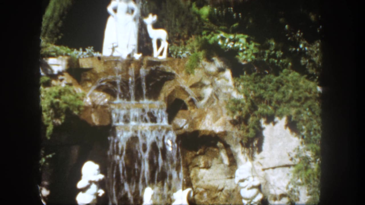 1962 Disneyland frame with the Snow White and deer statues above a tiered rock waterfall, partly framed by dark film borders.