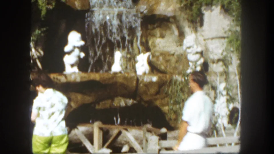 1962 Disneyland frame showing two park visitors near the Snow White waterfall display, with white figures, rockwork, and greenery behind them.