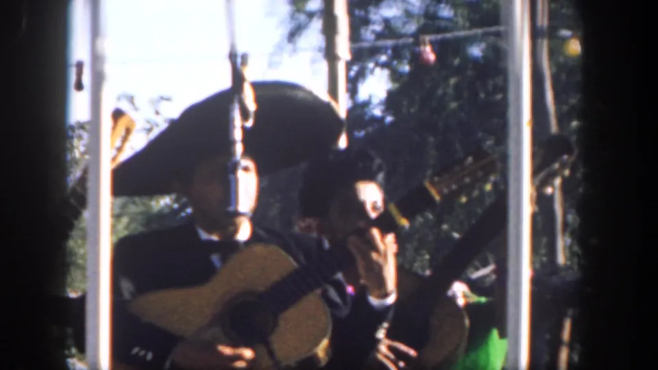 1962 Disneyland home-movie frame showing mariachi musicians in dark charro-style suits and sombreros playing guitars in sunlight.
