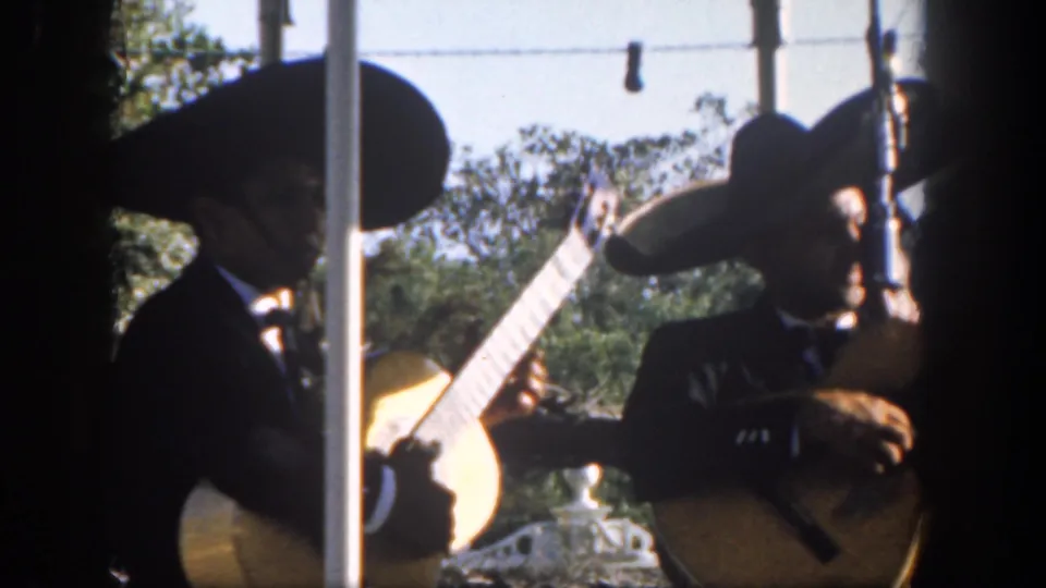 Visitor-shot 8mm frame from Disneyland shows mariachi musicians in sombreros playing guitars, with strong sunlight and foreground bars.