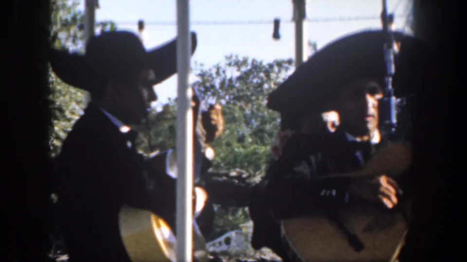 1962 Disneyland footage frame showing guitarists in sombreros and dark suits performing outdoors, partly shaded and partly silhouetted.