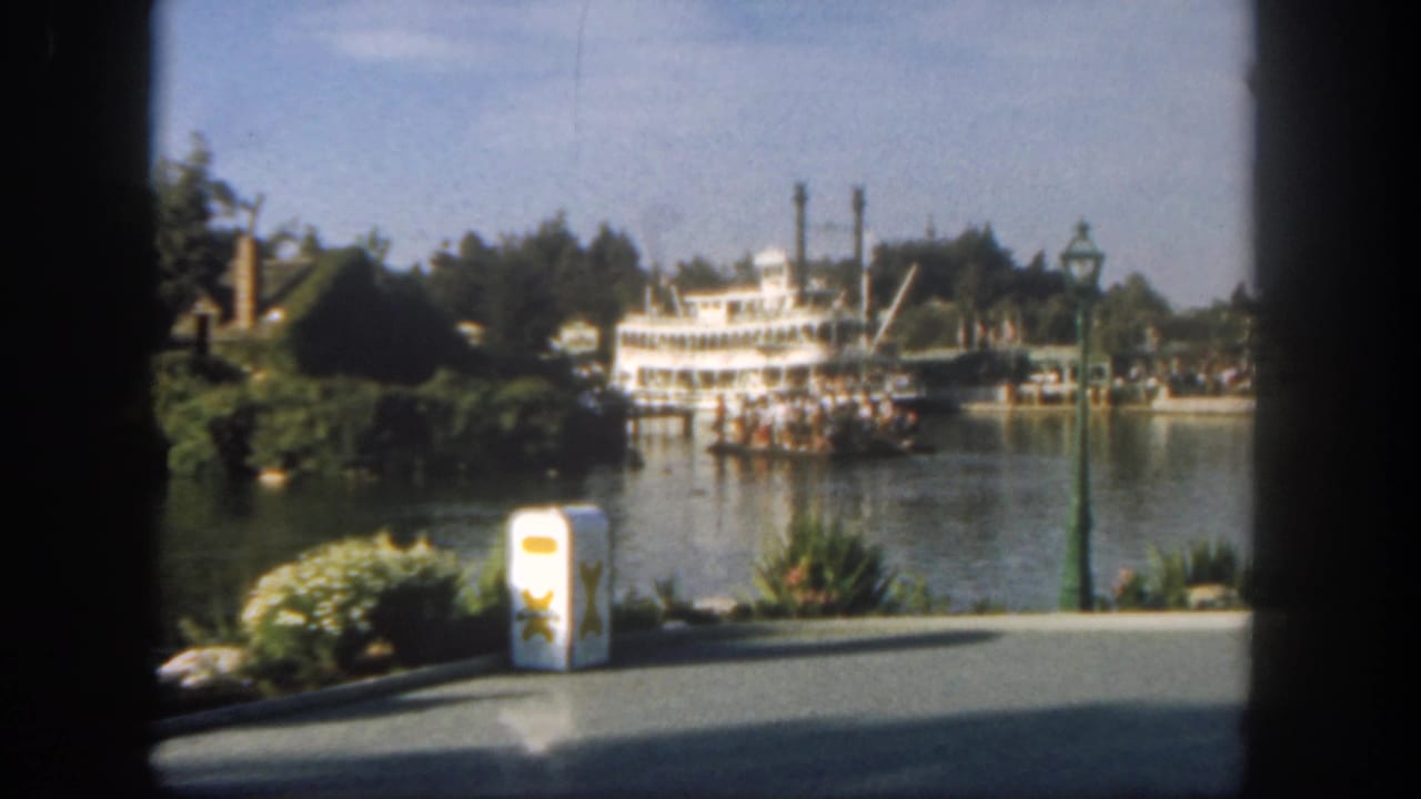 Steamboat and passengers on water with walkway and trash can in foreground.