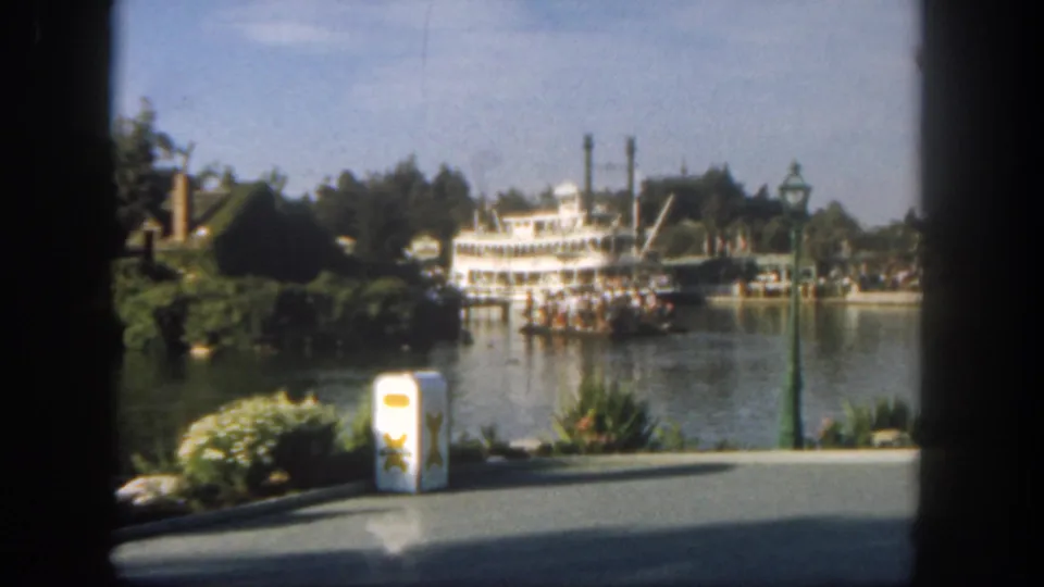 1962 Disneyland frame with a white steamboat carrying passengers, viewed over a paved path, landscaping, and a white trash can.