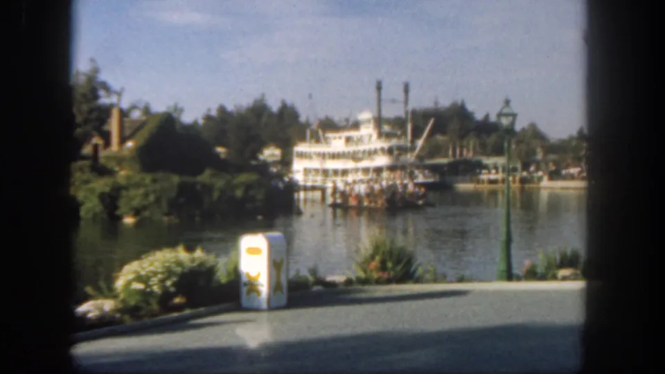 1962 Disneyland 8mm frame showing the steamboat slightly nearer the center, with shrubs, a trash can, water, and dark scan borders.