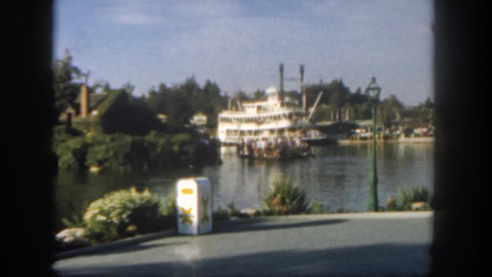 1962 Disneyland frame showing a white riverboat with passengers, calm water, foreground path, landscaping, and dark window-like edges.