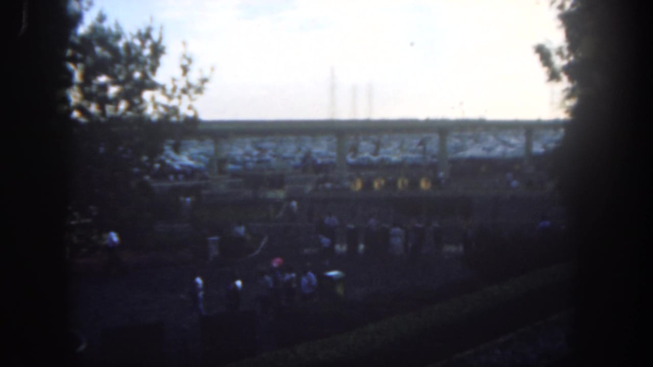 Crowds walk below a raised structure, seen through dark framing and silhouetted trees.