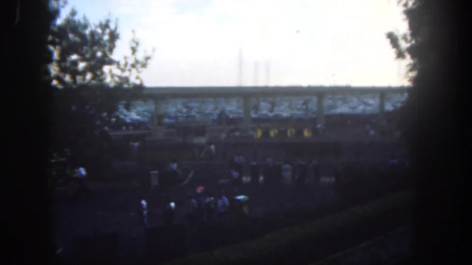 1962 Disneyland home-movie frame showing visitors moving below an elevated structure, with trees and dark frame edges around the view.