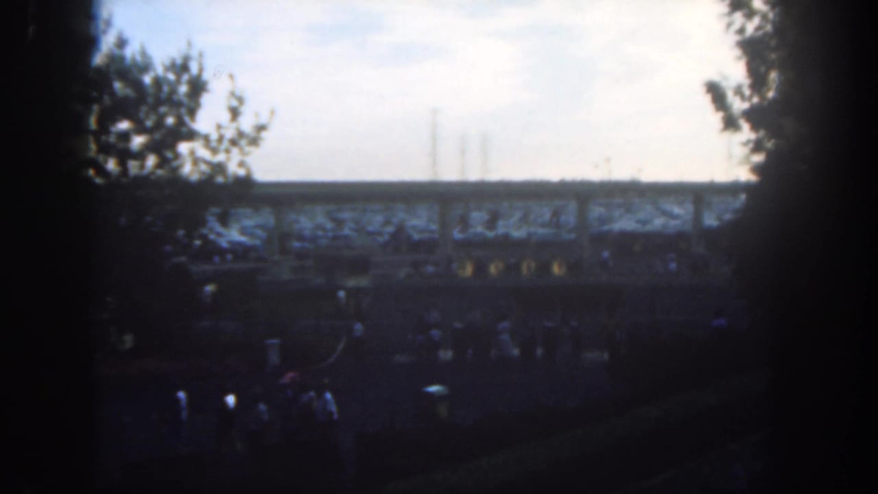 A dim view of park guests below a raised track, with trees and bright sky behind.