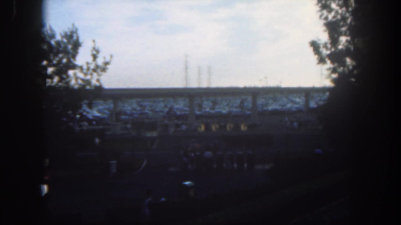1962 Disneyland 8mm frame with dark gate edges, trees, dusty ground, and distant rail or platform structures.