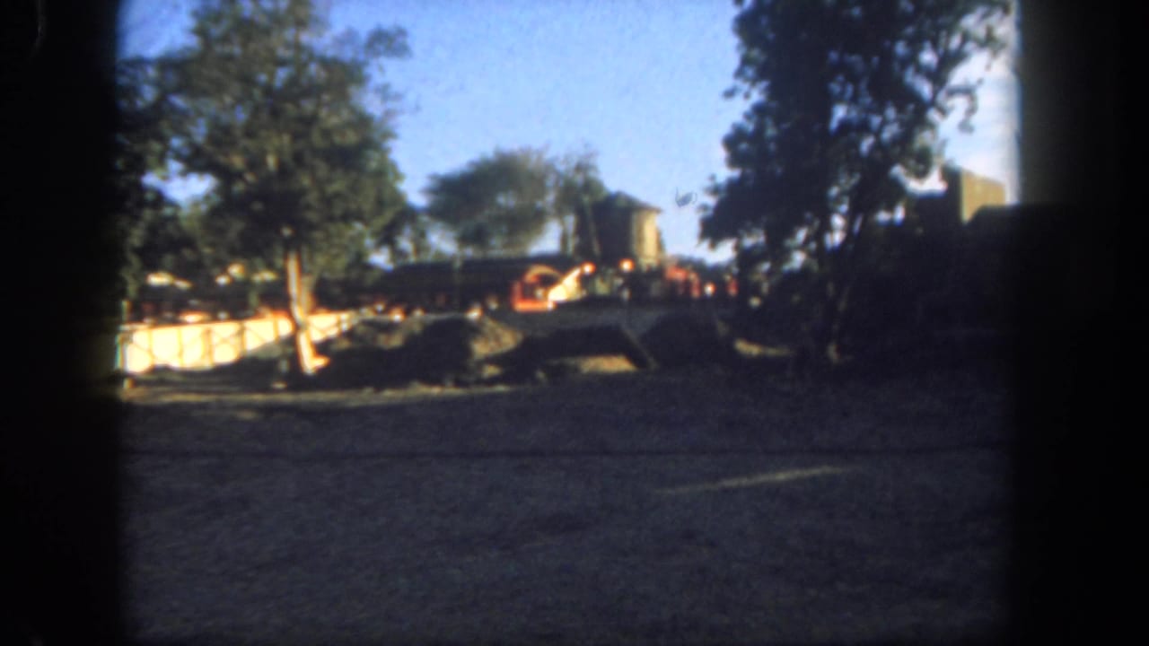 1962 Disneyland frame showing a sunlit dusty area, trees, rail tracks, and a small train or rail car near a themed structure.