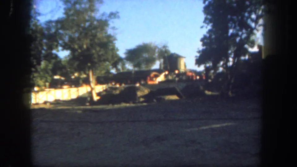 1962 Disneyland 8mm view of a dusty trackside area with trees, low barriers, and small rail equipment in bright sun.