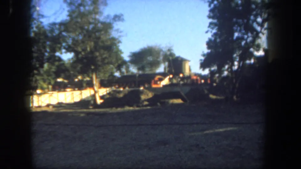 Final 1962 Disneyland frame with a dusty rail corridor, trees, low barriers, and a small train or rail car partly visible.