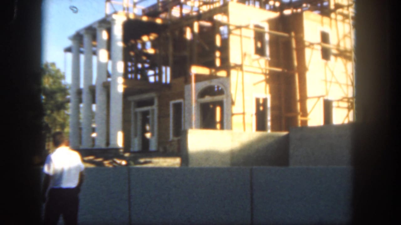 1962 Disneyland frame with a man walking past barriers before a white-columned building covered in scaffolding and wood framing.