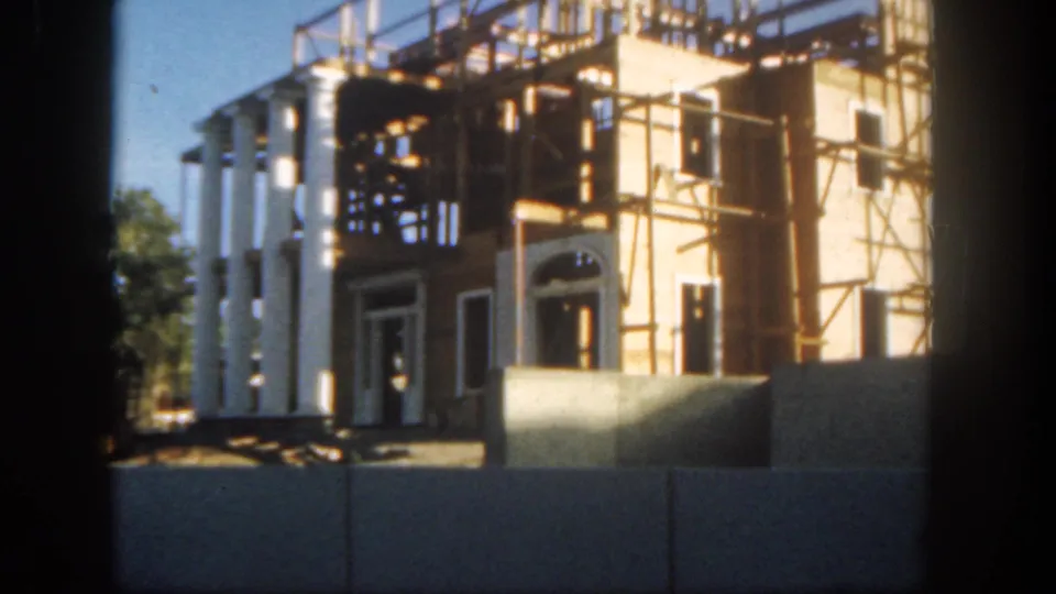 1962 Disneyland home-movie frame centered on a sunlit building under construction with white columns, scaffolding, and barriers.