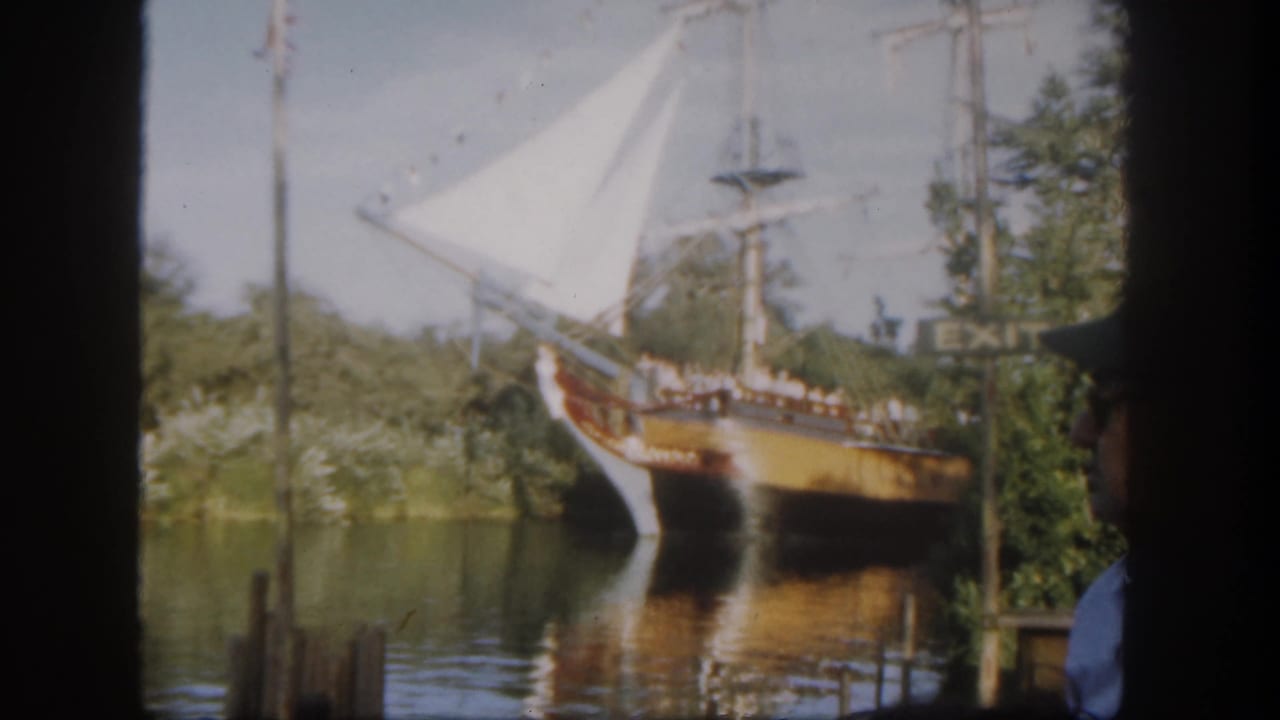 Wooden-style sailing ship with white sails on calm water, partly blocked by dark foreground edges.
