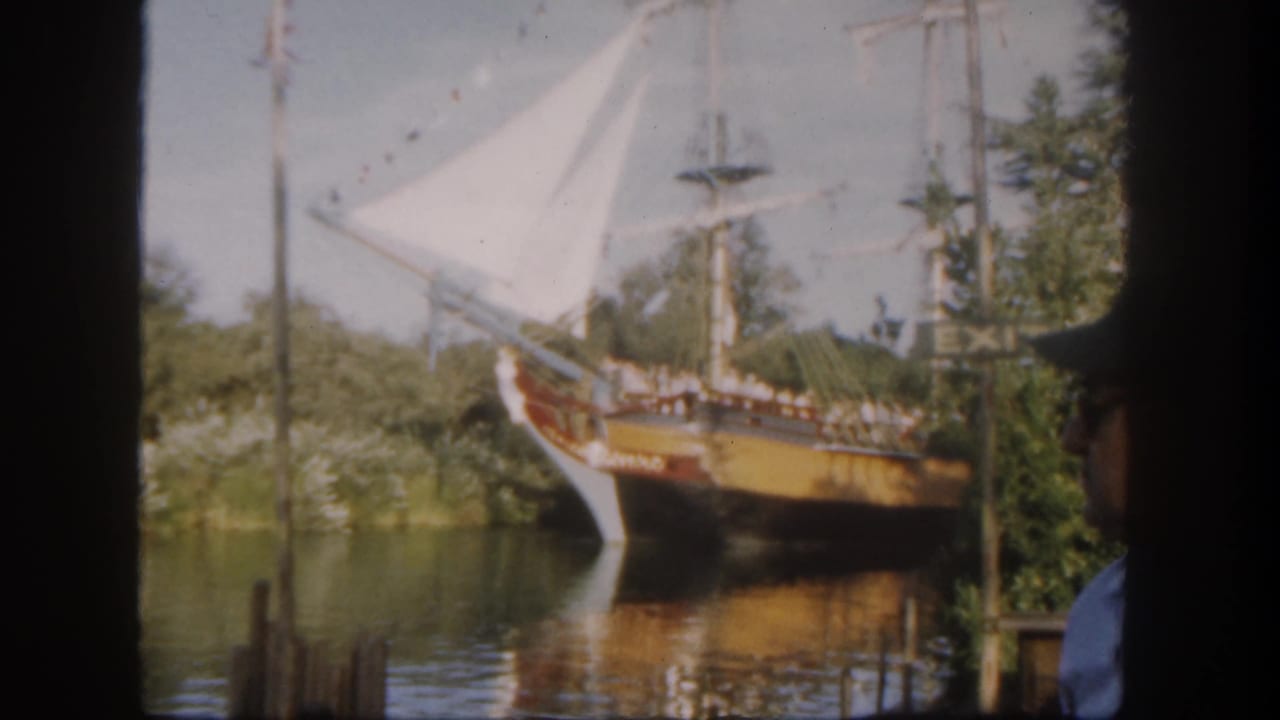Sailing ship passes near shoreline greenery with an EXIT sign and a partial man in a cap at right.