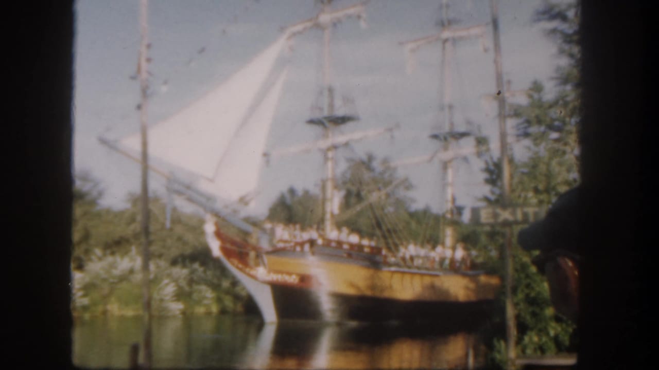 Wooden sailing ship with white sails on a waterway, seen through dark film borders.
