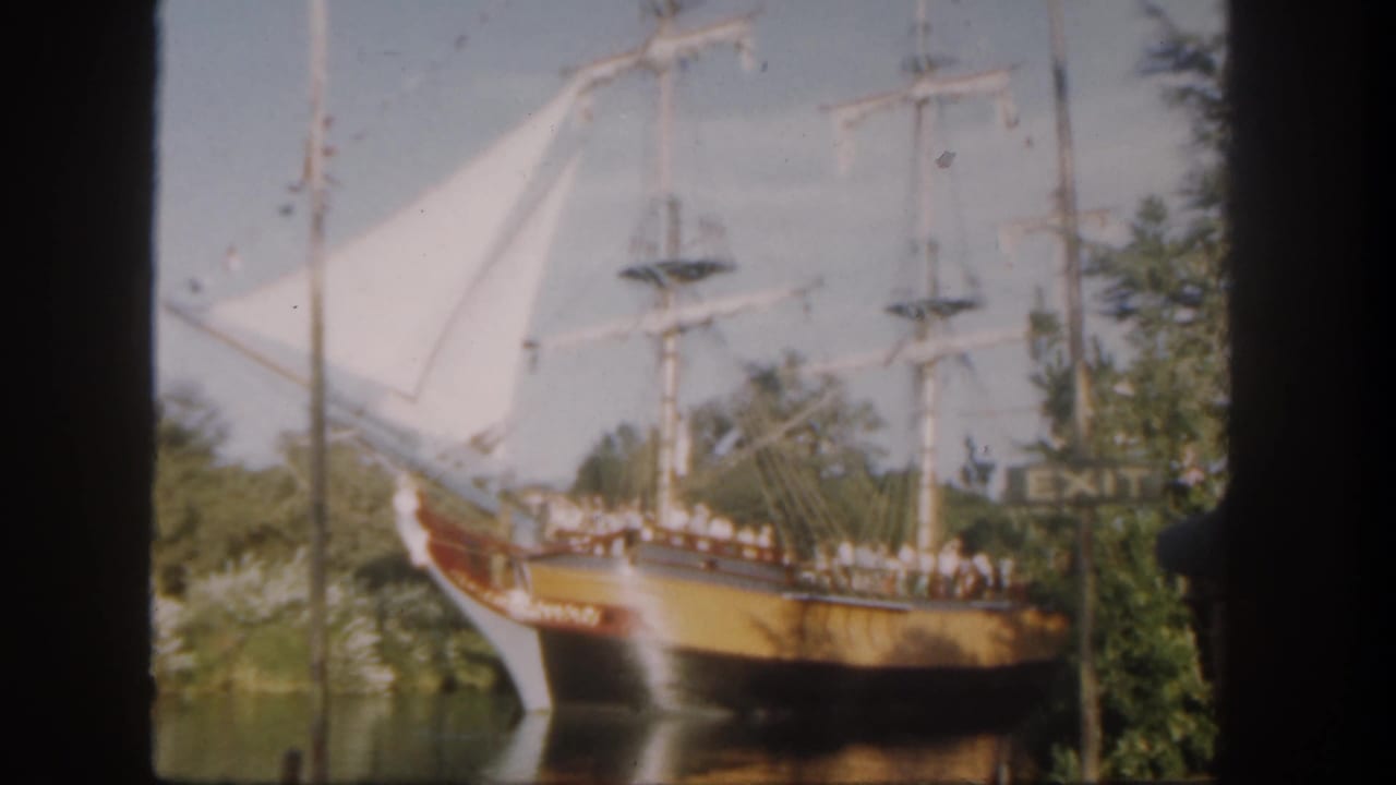 1962 Disneyland frame with a docked wooden sailing ship, white sails, visitors along the rail, trees, and a visible EXIT sign.