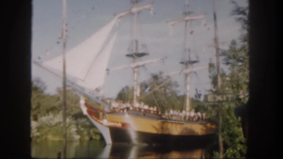 1962 Disneyland frame with a docked wooden sailing ship, white sails, visitors along the rail, trees, and a visible EXIT sign.