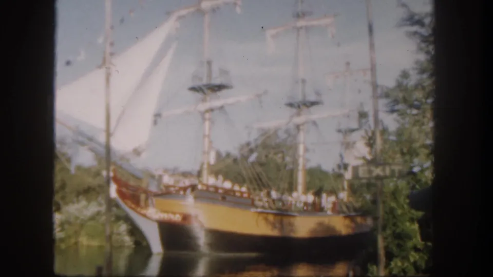1962 Disneyland frame of a wooden sailing ship attraction with passengers at the rail, white sails, trees, water, and an EXIT sign nearby.