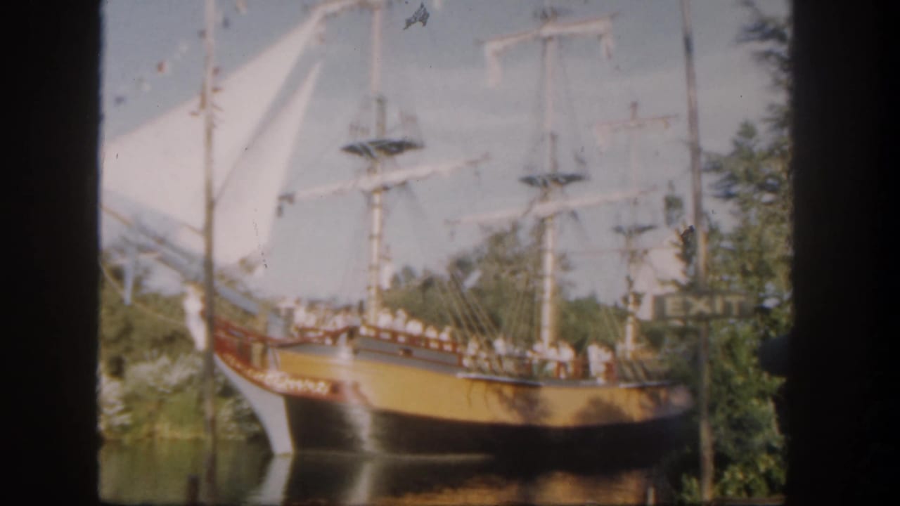 Replica sailing ship with white sails moves past trees and water.
