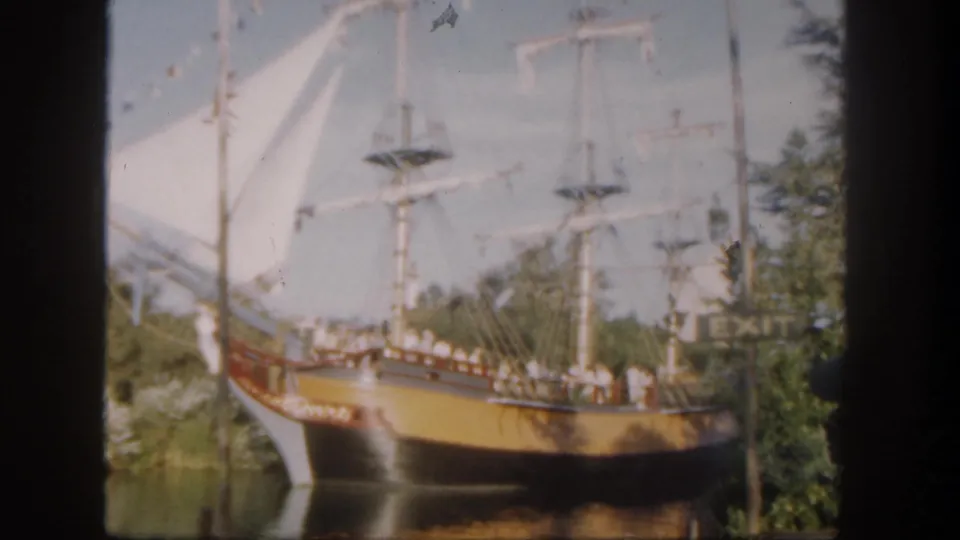 1962 Disneyland home-movie frame showing a colorful replica sailing ship with white sails, rigging, passengers, water, and trees.