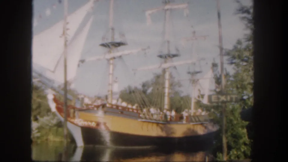 1962 Disneyland frame with a replica ship under white sails, visitors on deck, green trees, water, and an EXIT sign in the background.
