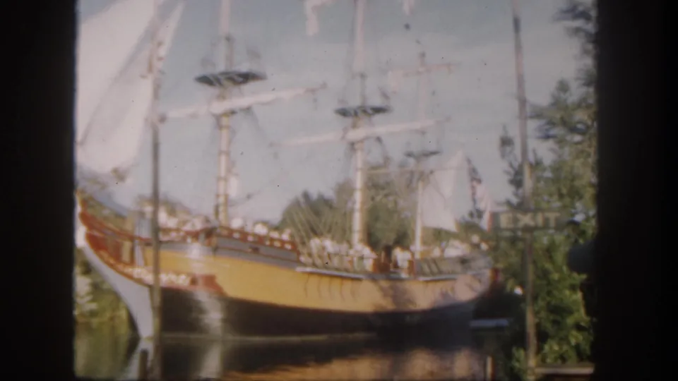 1962 Disneyland home-movie still showing the replica sailing ship beside trees and water, with passengers on deck and an EXIT sign behind it.